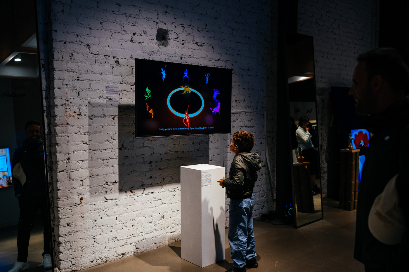 Child stands before a wall-mounted screen displaying colorful animated figures in a brick-walled gallery.