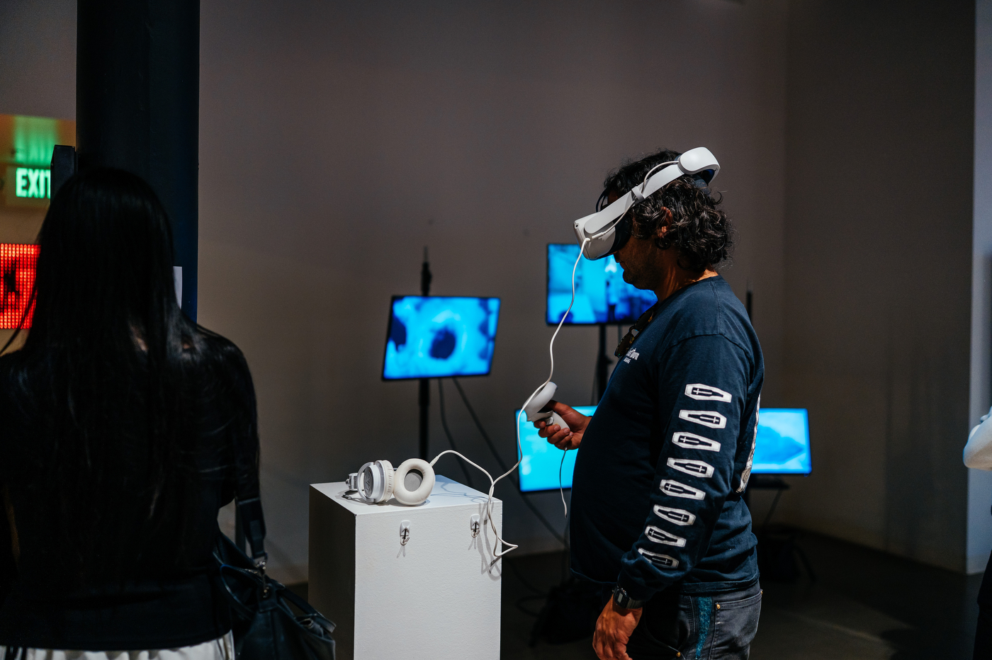 Visitor wearing a VR headset and holding controllers interacts with a digital exhibit, with a screen showing blue graphics in the background.
