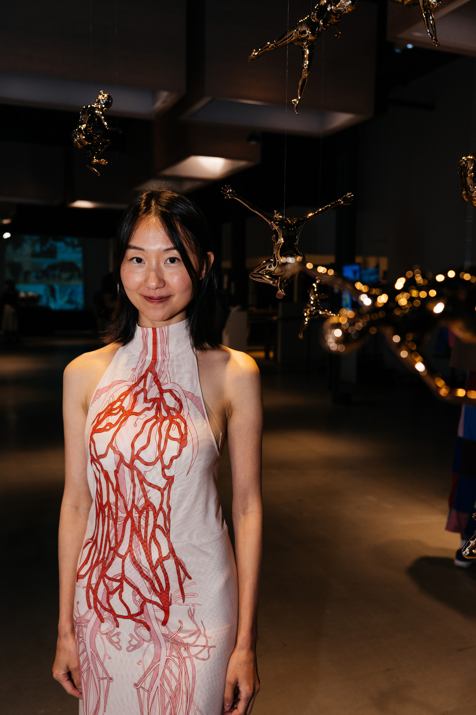 Woman wearing a white dress with a red vein-like pattern stands in a gallery space with hanging sculptural figures.