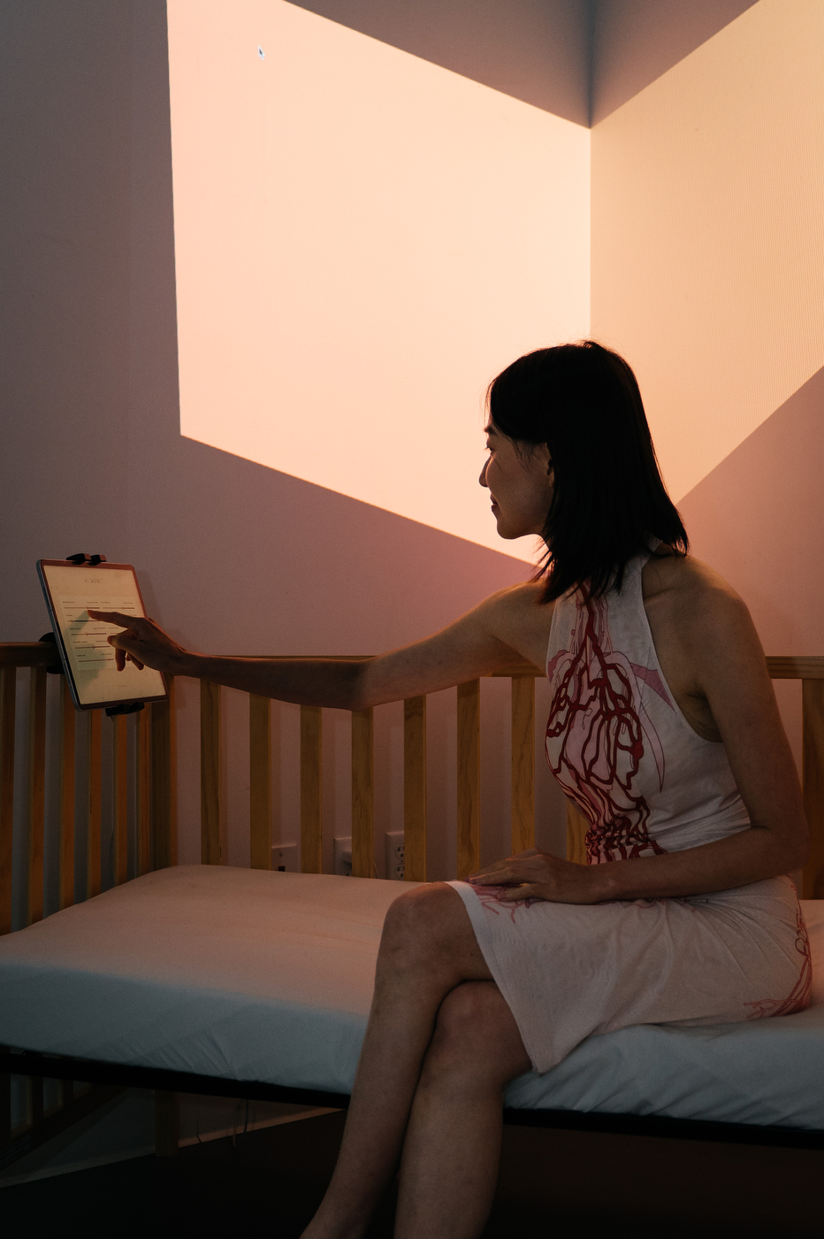 A person in a vein-patterned dress sits inside a crib-like installation, reaching toward a small tablet, lit by warm orange light against a triangular wall.