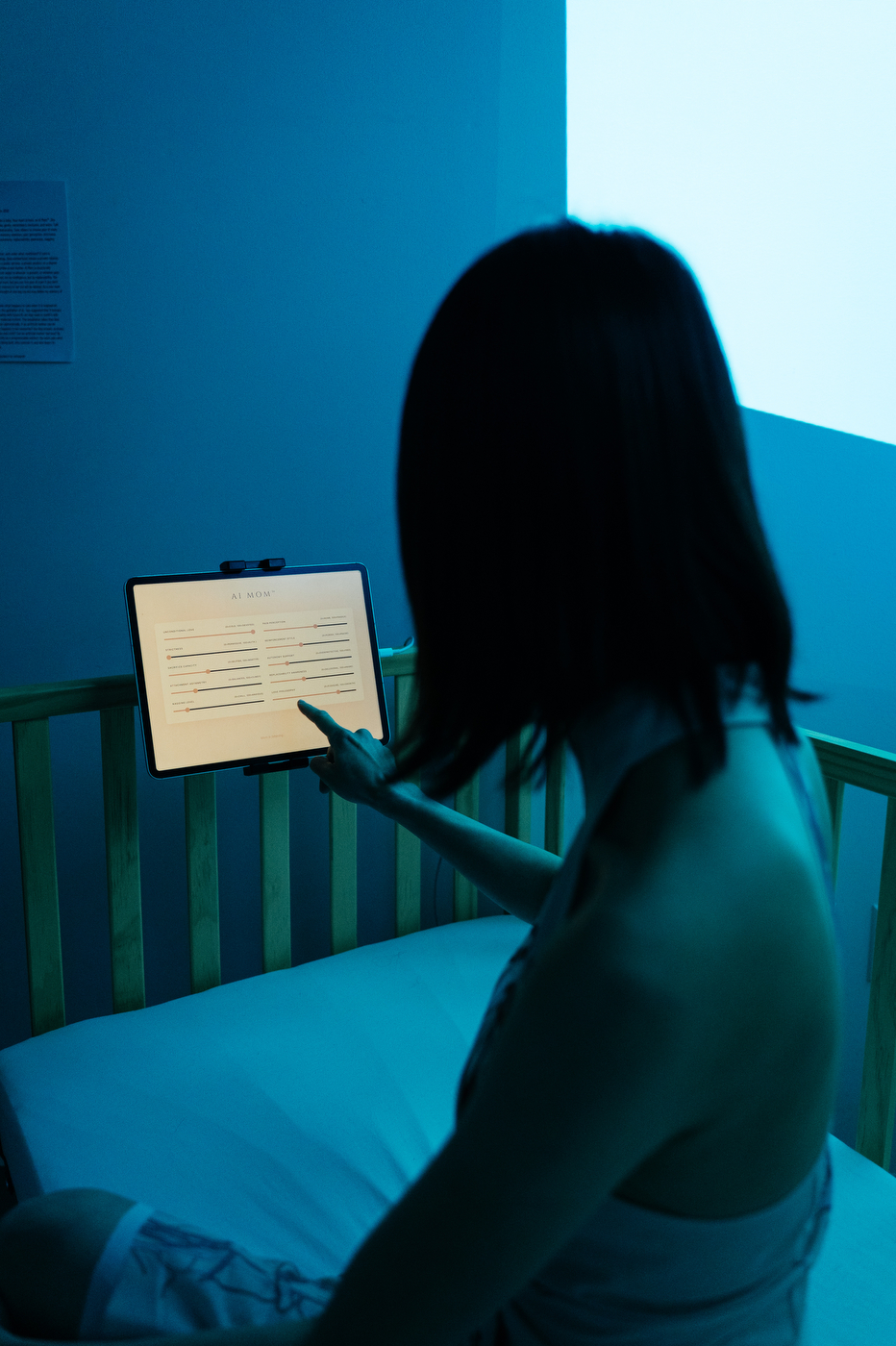 A person sits inside the same crib-like installation reaching toward a tablet, lit in cool blue light.