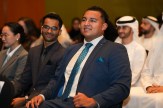 Vicent Armas seated in an audience, smiling, wearing a dark suit with a teal tie, among other attendees in business attire and traditional Gulf dress.