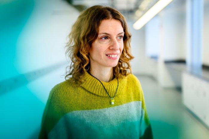 Dimitrova smiles slightly, looking off to the side, wearing a green sweater in a bright office hallway.