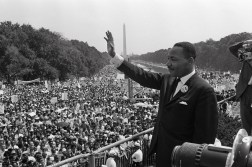 Martin Luther King Jr. raises his right hand while speaking from a podium crowded with microphones, addressing a dense crowd that stretches back toward the Washington Monument.