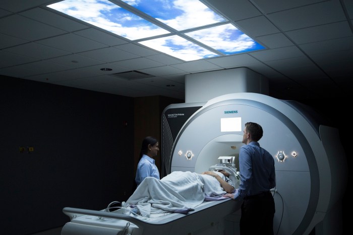 Two staff members standing on either side of an MRI scanner with a patient laying on a table in between them, covered with a blanket.