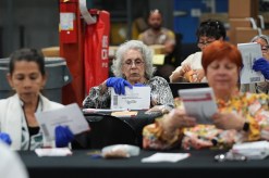 Workers wearing blue gloves sit at tables examining ballot envelopes and papers.