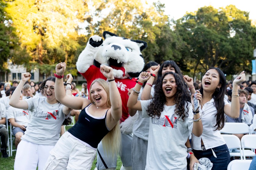 Students cheer with arms raised alongside Northeastern's husky mascot at an outdoor welcome event, with a large crowd seated behind them.