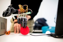 Robotic prosthetic hand with exposed wiring grips a red disc, with a laptop and small objects nearby on a desk.