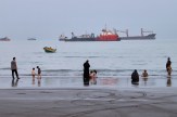 Families wade at the shoreline while several large cargo ships sit anchored in the hazy water behind them.