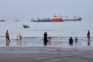 Families wade at the shoreline while several large cargo ships sit anchored in the hazy water behind them.