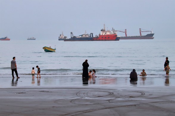 Families wade at the shoreline while several large cargo ships sit anchored in the hazy water behind them.