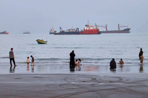 Families wade at the shoreline while several large cargo ships sit anchored in the hazy water behind them.