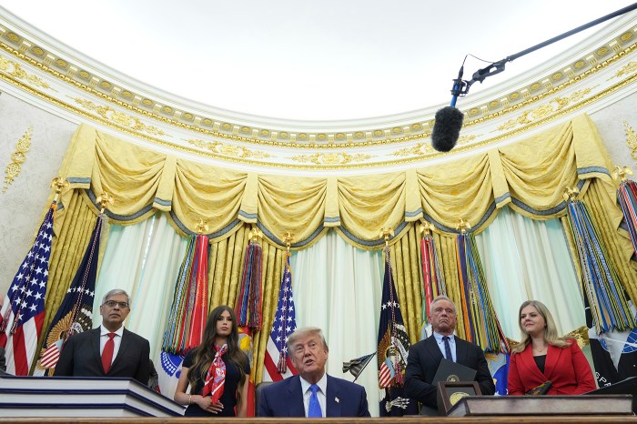 Trump seated at a desk with five officials standing behind him, in front of the Oval Office's gold curtains and flags, a boom microphone extending overhead.