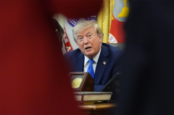 President Trump seated at the Resolute Desk, seen through the shoulders of two people in the foreground, mid-speech.