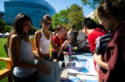 Students browse a row of club tables set up on the quad, talking with members staffing the booths under a sunny sky.