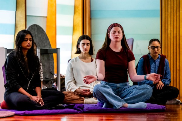 Students sit cross-legged in a meditation pose with eyes closed, hands resting on their knees in mudra position.