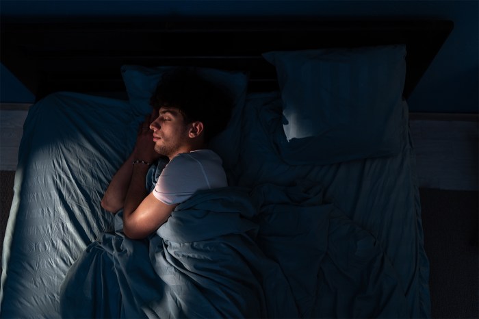 A young man asleep in bed, curled under blankets in dim, blue-toned light.