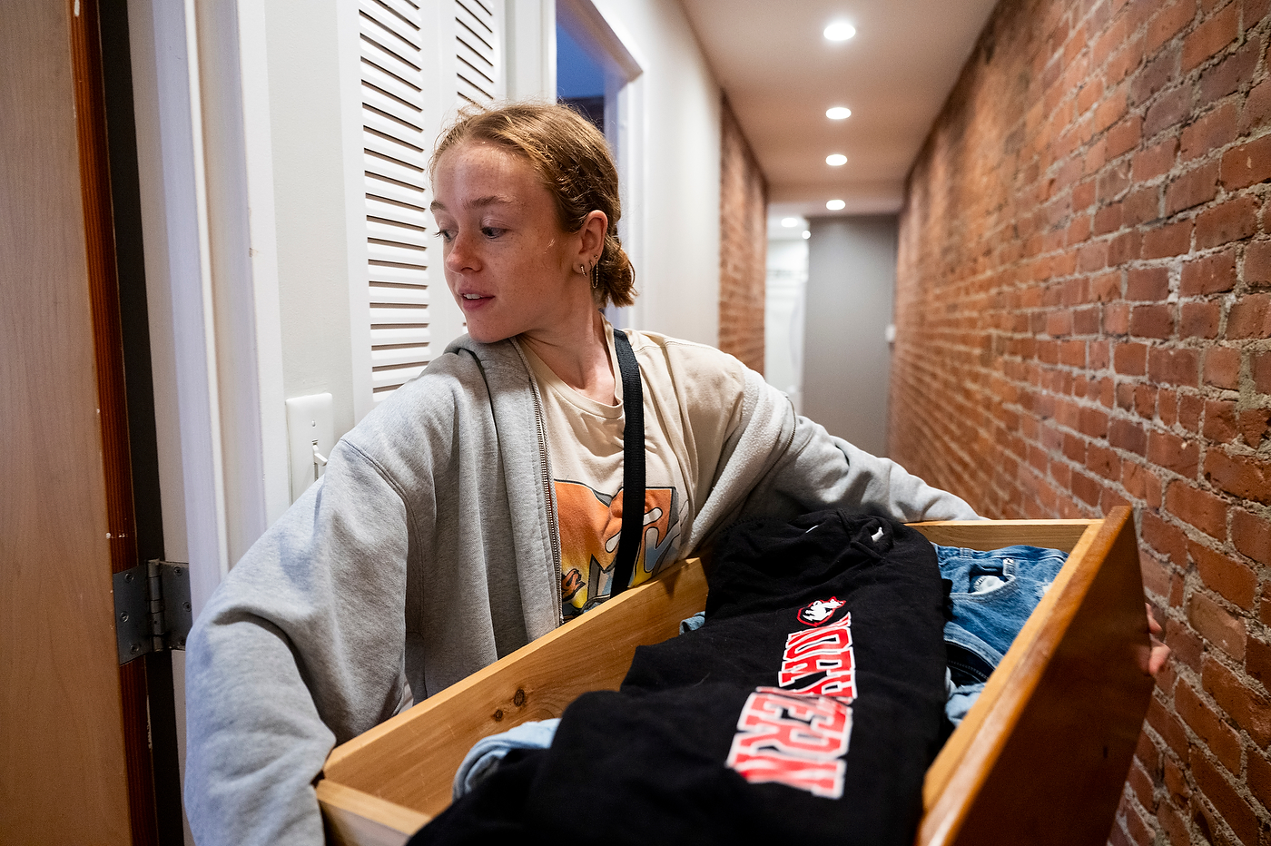 Caroline Scalzulli carries a wooden drawer full of belongings through a doorway in a brick-lined hallway. 