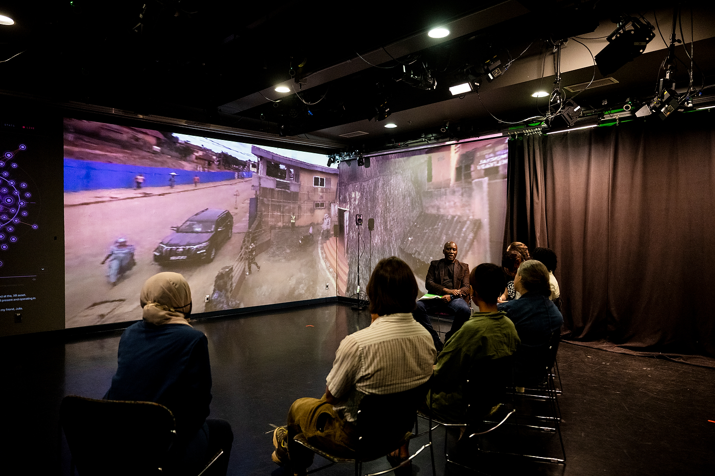 Students seated in a darkened studio, viewing a large wraparound projection of a street scene with a car and figures.