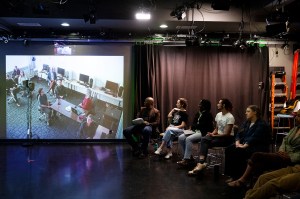 Students seated in a row of stools face a large projection screen showing a video call with collaborators, inside a darkened studio space with theater lighting rigs overhead.