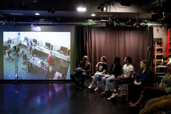 Students seated in a row of stools face a large projection screen showing a video call with collaborators, inside a darkened studio space with theater lighting rigs overhead.