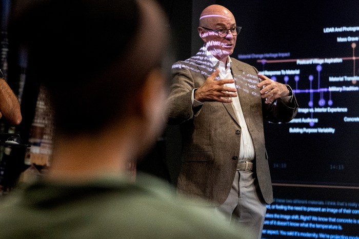 President Aoun gestures while speaking to a group, with projected light patterns across his face and a data diagram visible on screen behind him. 