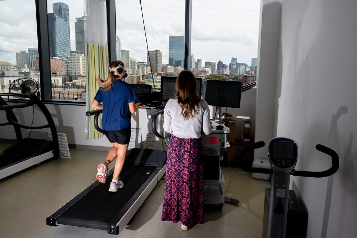 Smith corre en una cinta de correr frente a una ventana del piso al techo con vista al horizonte de Boston mientras Carey monitorea los datos en una estación de trabajo cercana.