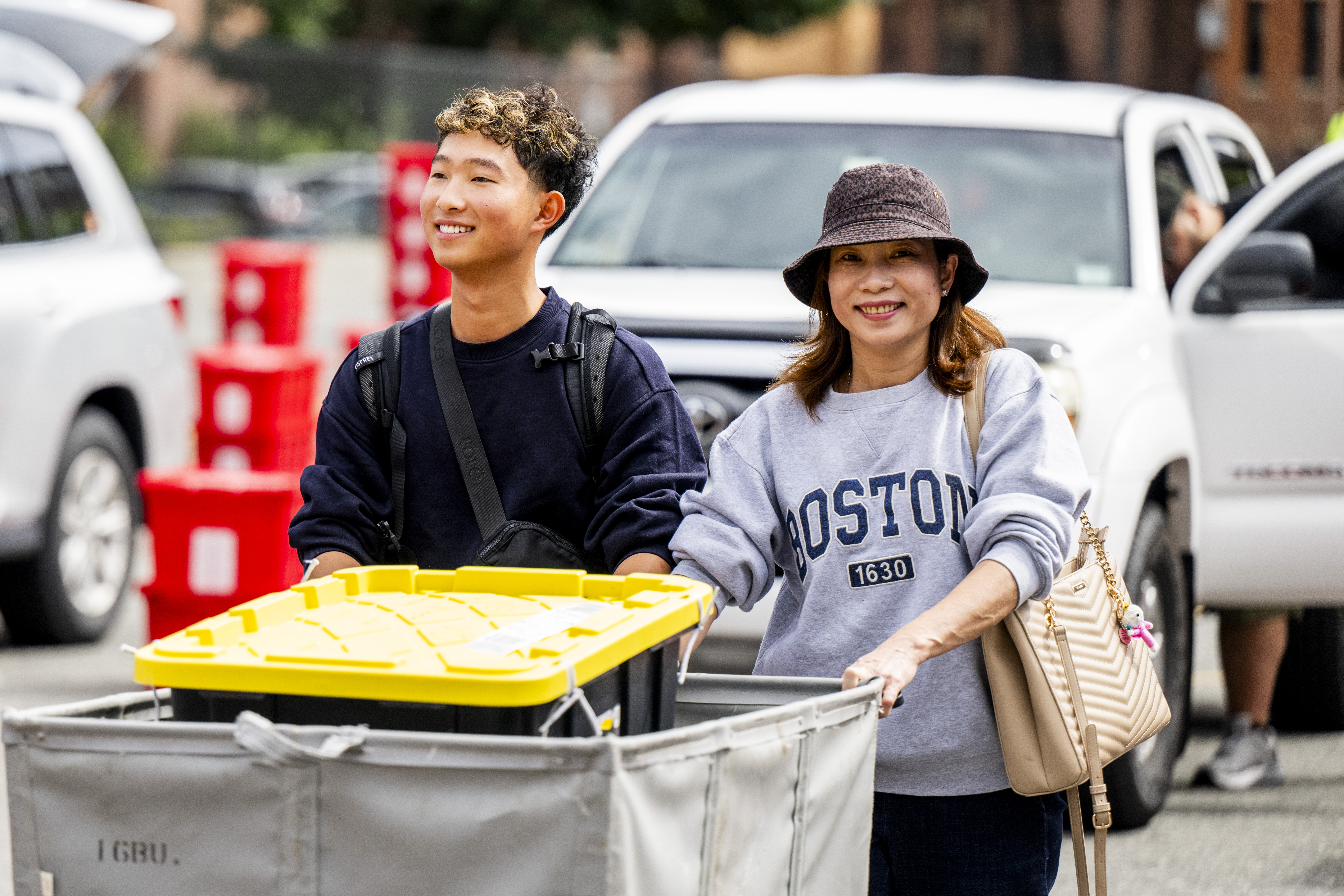 A woman in a bucket hat walking next to a young man wheeling a move-in cart in front of them. Behind them are parked cars.