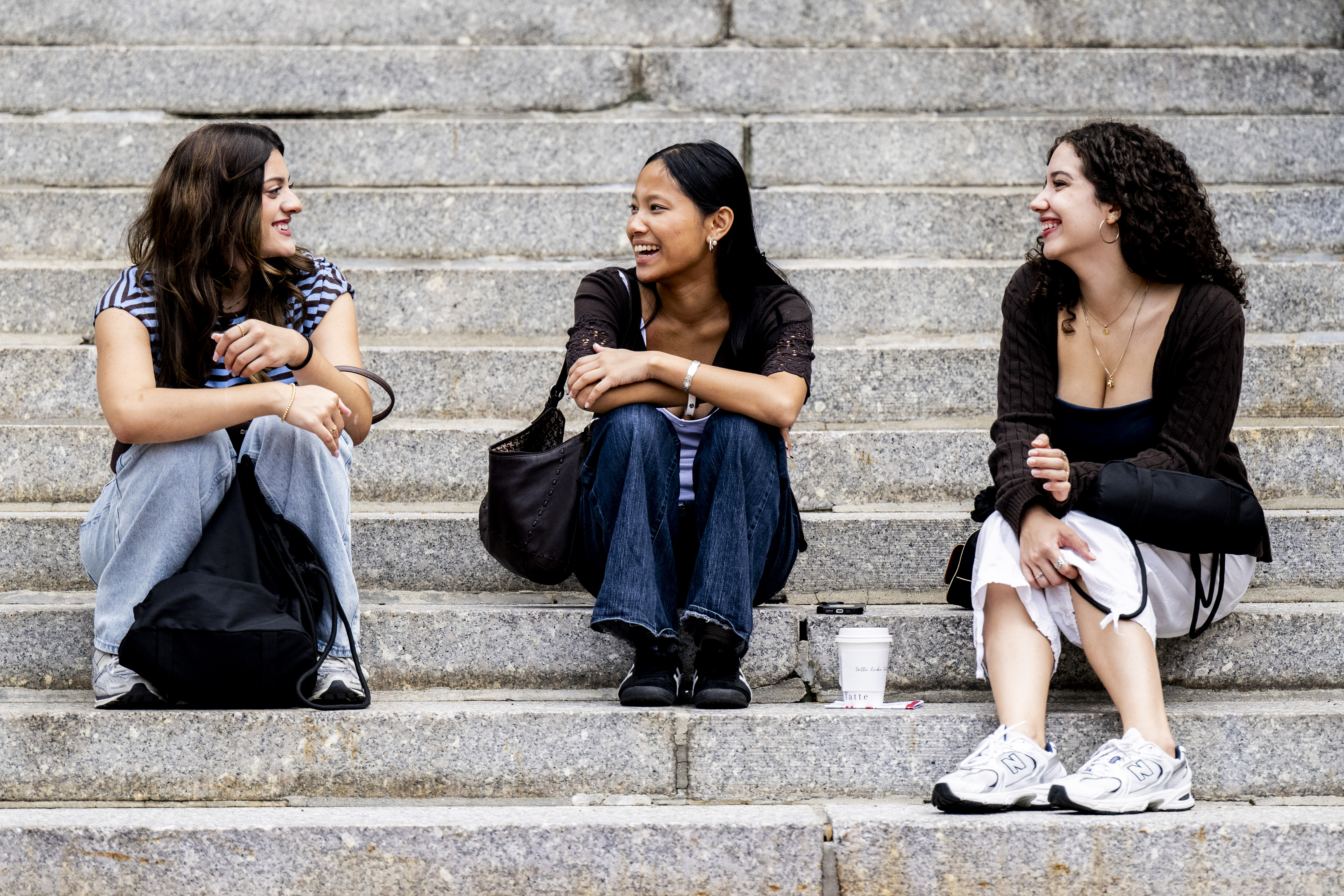 Three students sit and talk on outdoor stone steps, backpacks and drinks beside them.