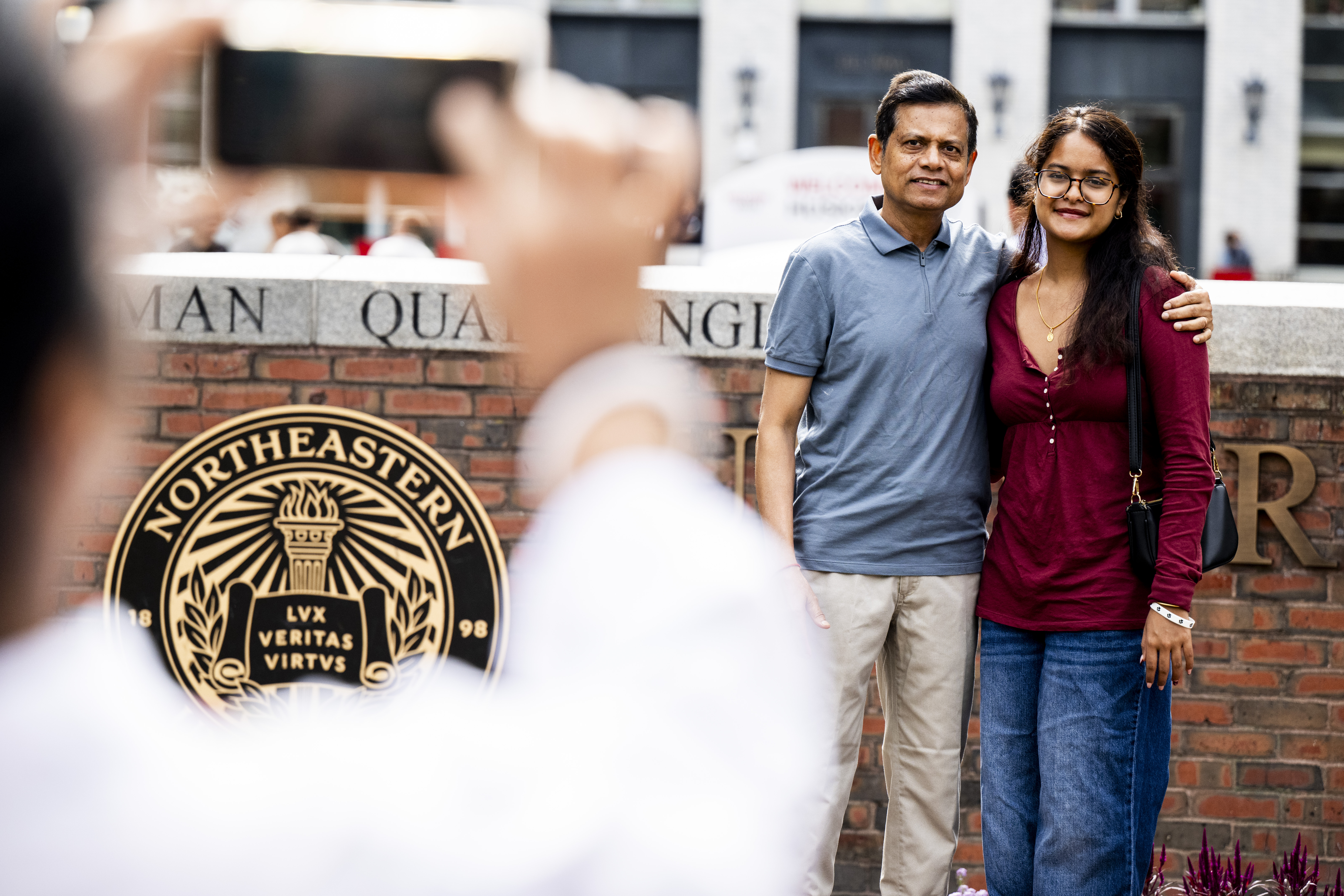 A man and a young woman pose with arms around each other in front of the Northeastern seal on a stone wall, while a bystander takes their photo in the foreground.