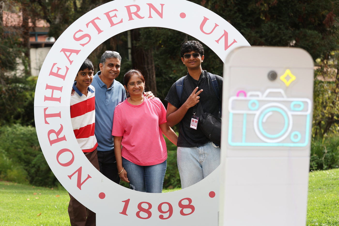 A family poses inside a large photo-op cutout shaped like a Northeastern seal with a phone camera graphic.