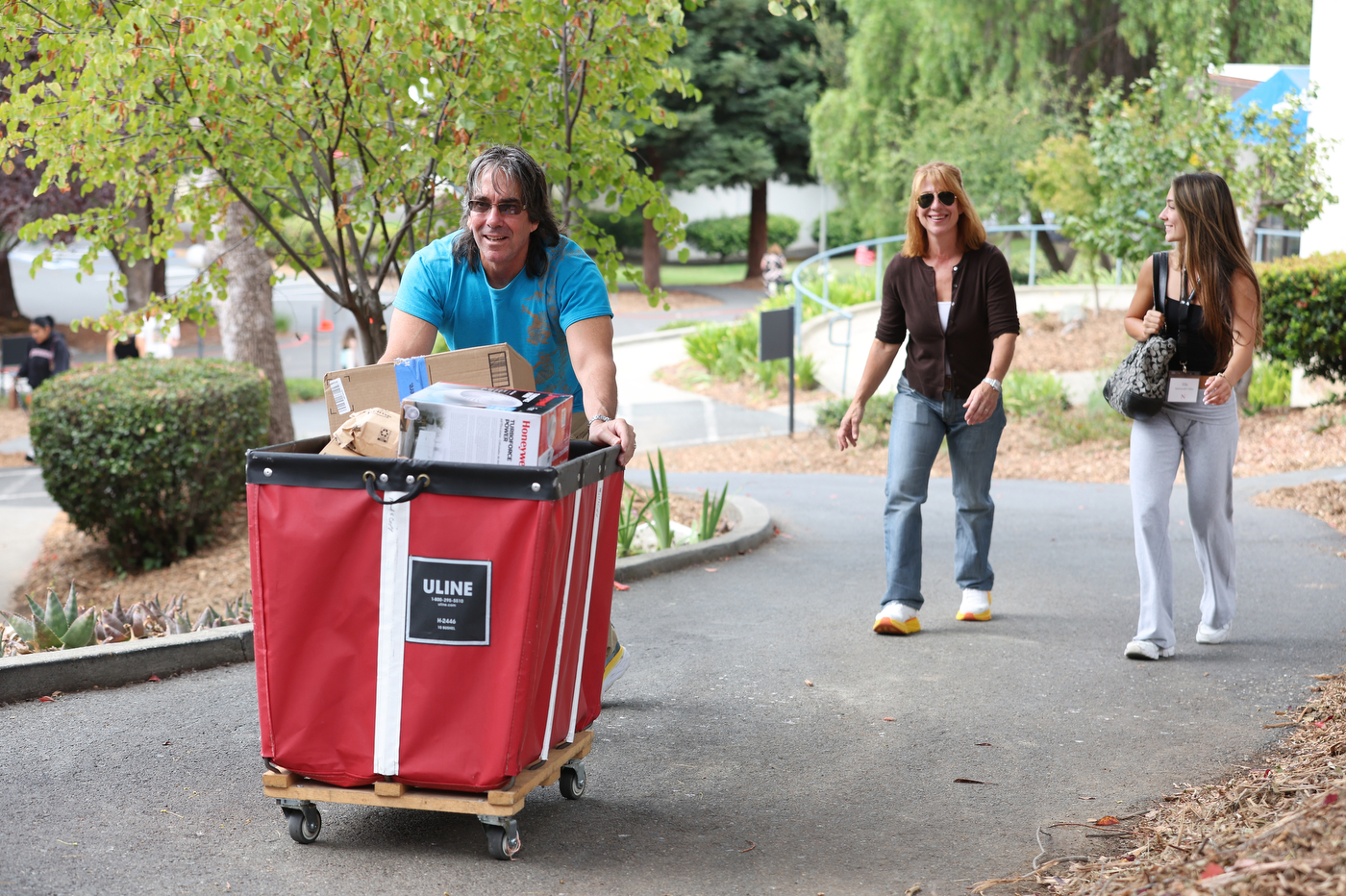 A man pushes a cart of boxes along a campus path while two women walk alongside him.