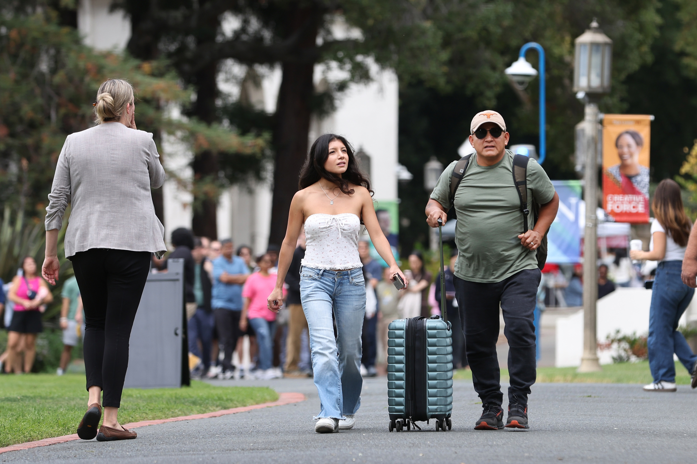 A family walks toward a building on campus, one pulling a rolling suitcase, with a crowd gathered near the entrance.