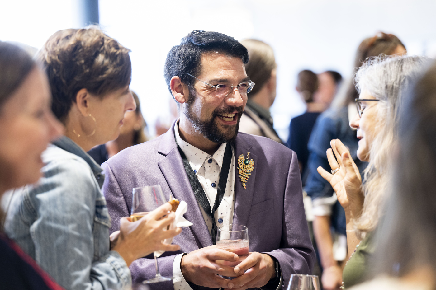 A man in a lavender blazer holding a drink and appetizer, laughing in conversation with other attendees.