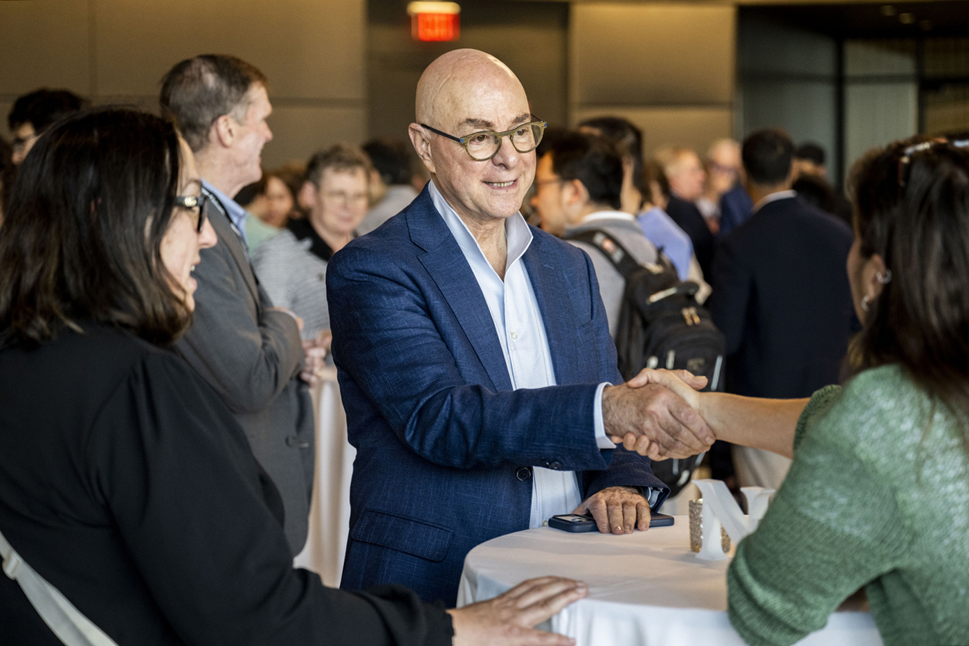 President Joseph E. Aoun shaking hands with an attendee at a standing-room reception, surrounded by other guests in conversation.