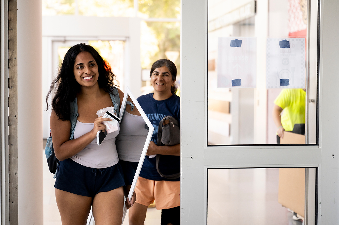 Two students smile while carrying a mirror and belongings into a residence hall.