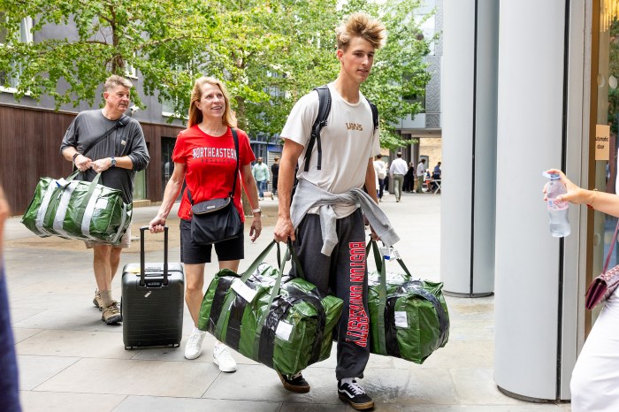 A student carries two large duffel bags while walking with family members, one wearing a Northeastern Huskies t-shirt, arriving at a building entrance on the London campus.