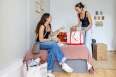 Two students unpacking in a dorm room, one sitting on a bed talking while the other stands unpacking a Northeastern tote bag, with moving boxes and framed photos on the wall.