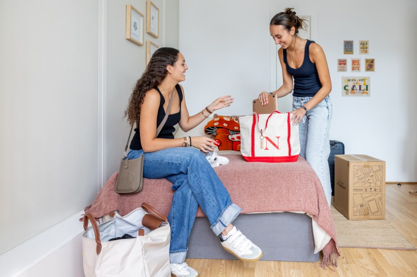 Two students unpacking in a dorm room, one sitting on a bed talking while the other stands unpacking a Northeastern tote bag, with moving boxes and framed photos on the wall.