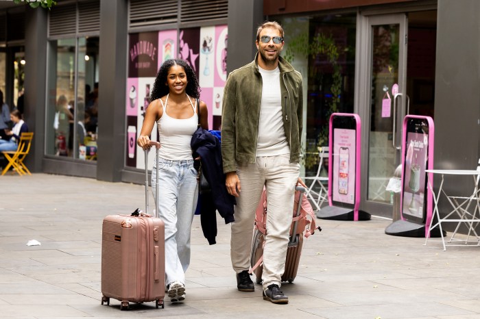 A student pulling a rolling suitcase down a city sidewalk beside an older man, passing storefronts near Northeastern's London campus.