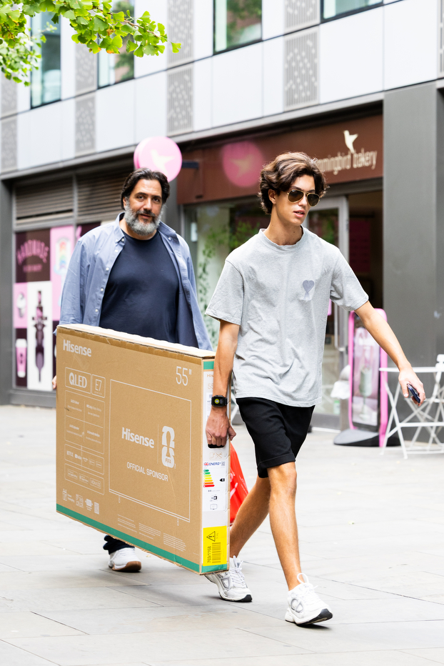 A student and a man carrying a large TV box together down a city sidewalk.