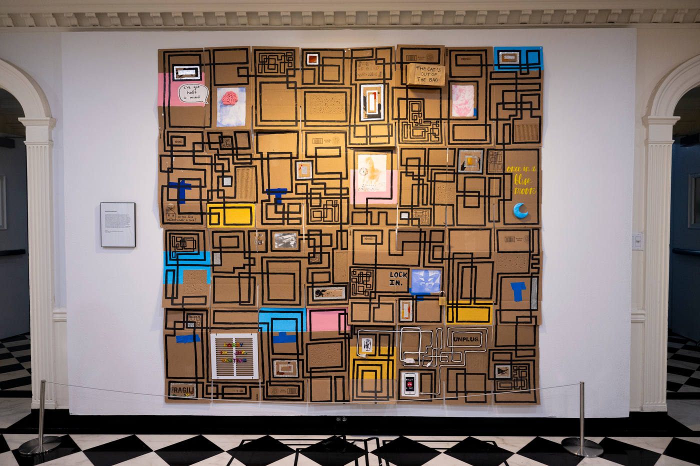 Large mixed-media wall installation made from cardboard panels covered in black geometric lines, with smaller colorful artworks, handwritten text and abstract designs arranged throughout.