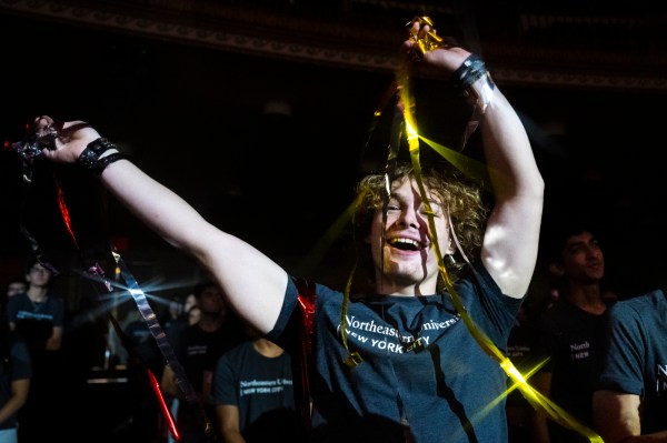 A Northeastern University student in a gray T-shirt raises both arms and smiles while holding metallic gold and red streamers during a lively event at The Town Hall in New York City. Other students stand in the dimly lit auditorium behind them.