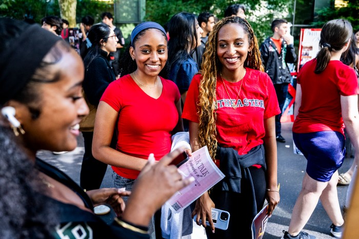 Three students smile and talk together in a crowd, one holding a flyer.