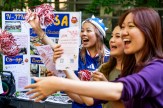 Students cheering and waving red and white pom-poms while reaching toward the camera in front of a Japanese Student Association club table.