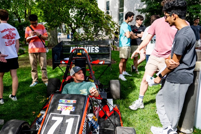 A student sits in a small open-cockpit race car on the grass, as other students look on and talk nearby.