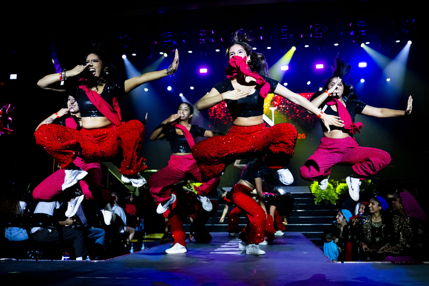 Dancers in red pants and black tops perform mid-jump on stage during the convocation program.