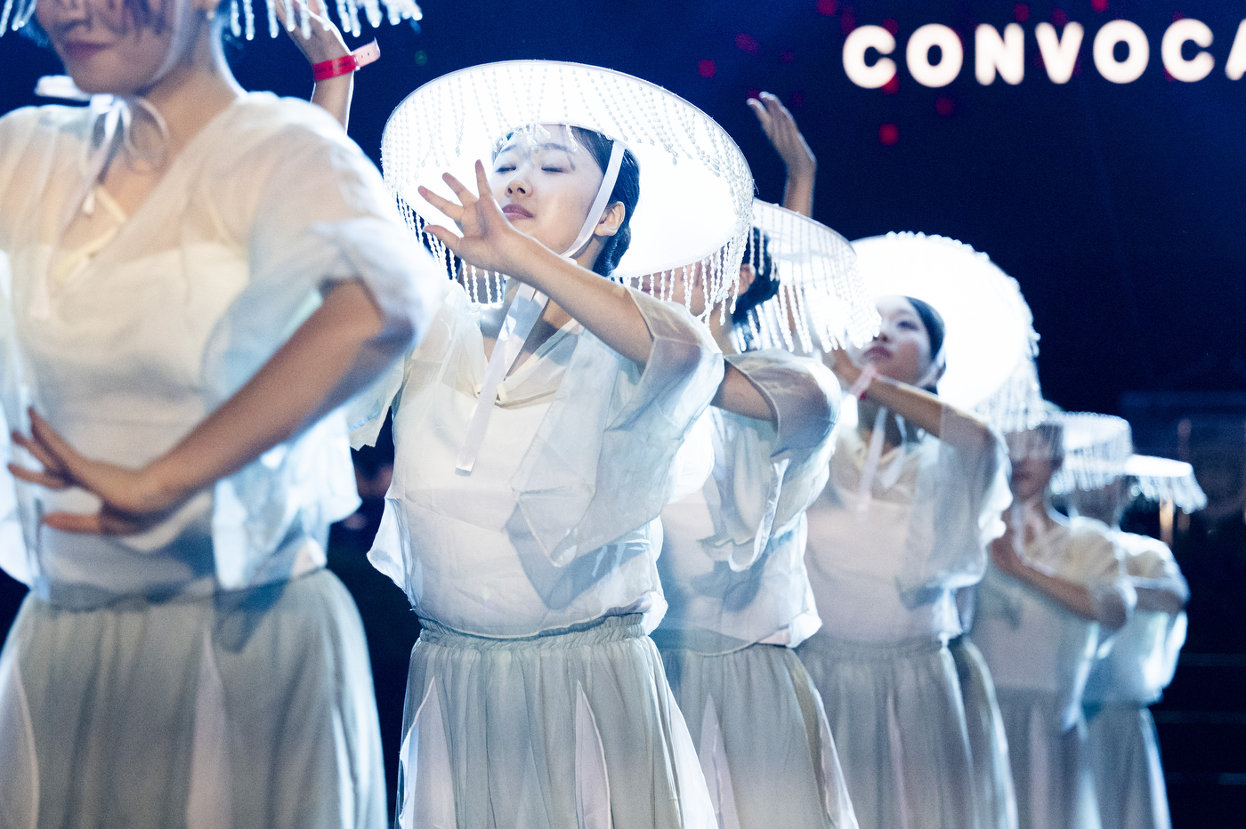 Performers in white feathered headpieces and costumes on stage during the convocation program.