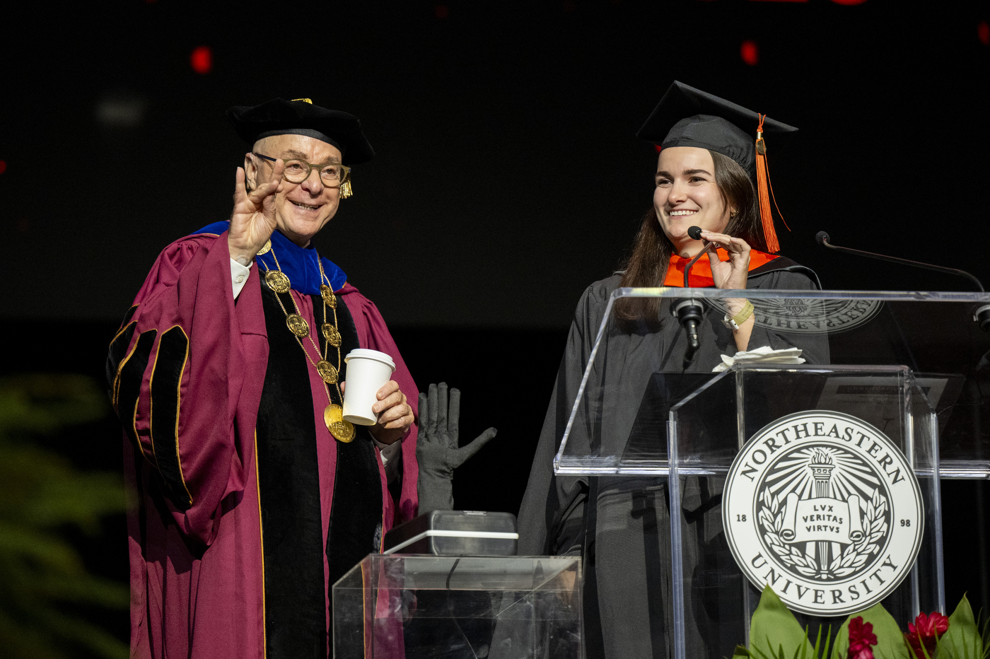 President Joseph E. Aoun on stage in academic regalia, holding up one hand with thumb and forefinger touching and holding a coffee cup in the other hand.