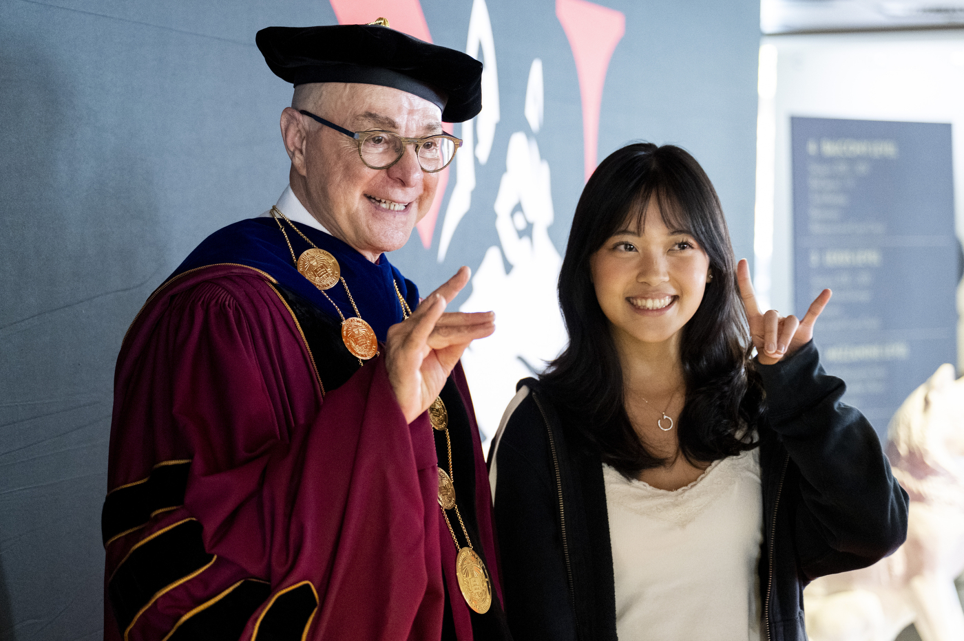 President Aoun in regalia poses with a student making a hand gesture.